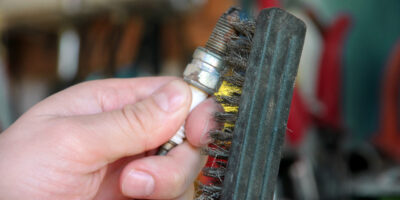 A person using a metal brush to clean a dirty spark plug. The background is out of focus but appears to be a garage.