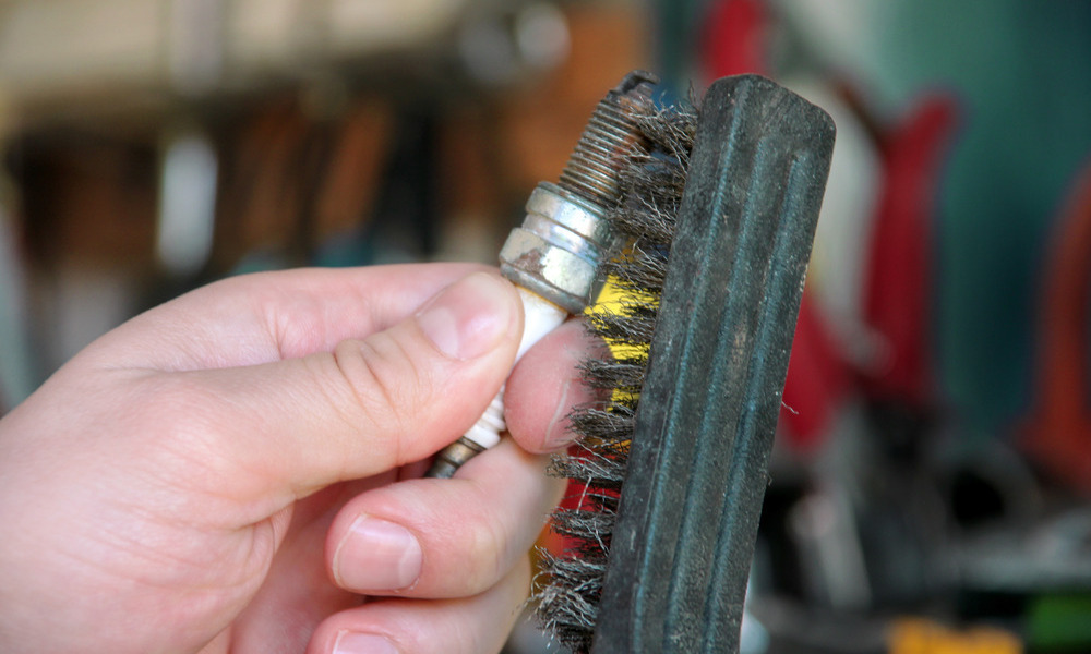 A person using a metal brush to clean a dirty spark plug. The background is out of focus but appears to be a garage.