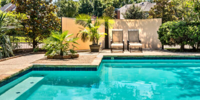 Closeup of a luxurious backyard pool with bright turquoise water next to a concrete patio with chairs and plants.