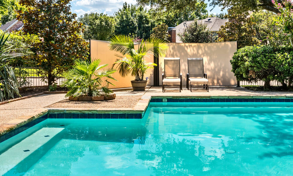 Closeup of a luxurious backyard pool with bright turquoise water next to a concrete patio with chairs and plants.