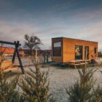 A tiny wooden home with large windows parked on a camping site with a picnic table and swinging bench next to it.