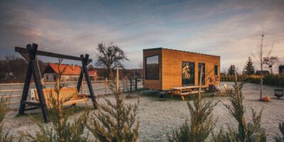 A tiny wooden home with large windows parked on a camping site with a picnic table and swinging bench next to it.