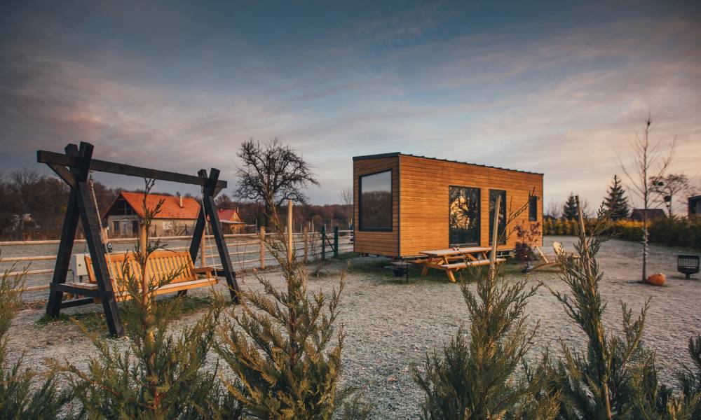 A tiny wooden home with large windows parked on a camping site with a picnic table and swinging bench next to it.