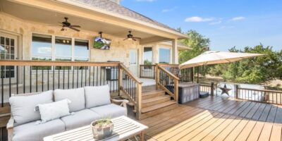 A backyard deck with a wooden table and a couch with gray cushions. It is a bright, partly cloudy day.