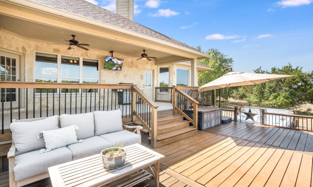 A backyard deck with a wooden table and a couch with gray cushions. It is a bright, partly cloudy day.