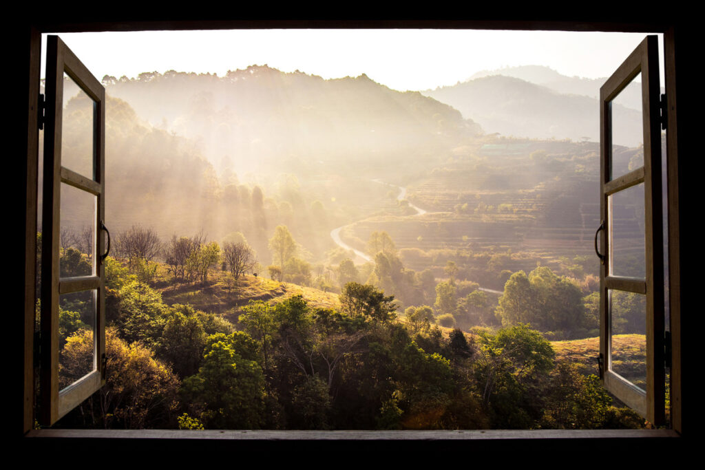 An open window looks out over an expansive outdoor area. There are rolling hills, lines of trees, and a winding road in the distance.