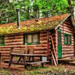 A rustic log cabin nestled in the woods surrounded by trees with a metal chimney and a picnic table next to it.