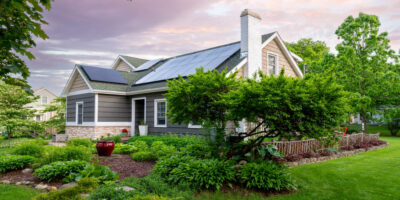 A small gray and beige home surrounded by trees features a set of solar power panels across its rooftop.