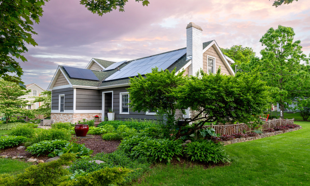 A small gray and beige home surrounded by trees features a set of solar power panels across its rooftop.