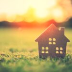 A small, wooden model of a house sitting on a patch of green grass with a sun setting in the background.