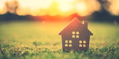 A small, wooden model of a house sitting on a patch of green grass with a sun setting in the background.