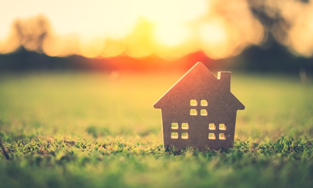 A small, wooden model of a house sitting on a patch of green grass with a sun setting in the background.